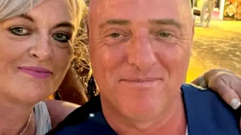 A "selfie" picture of McDonald and Mel on a beach. She has short blonde hair and is wearing pink lipstick. She has her arm around him and he is wearing a white t-shirt and blue suit waistcoat. They're both smiling.