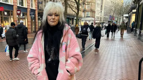 Sophie Wills stands on a Birmingham street with the top half of her hair dyed blonde and the other half black, wearing a pink cosy jacket