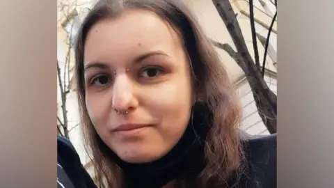 Family A woman with long brown hair, a brow piercing and a nose piercing, smiles at the camera. Trees without leaves are visible behind her.