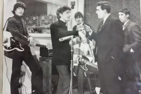 Johnny Haynes Five members of a band, two with guitars, look towards a man with a suit and tie