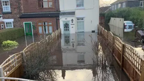 Polly Pee A view of the rear of a row of terraced houses which all have rear gardens submerged in brown, muddy flood water