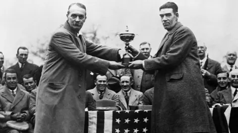 Getty Images Samuel Ryder, with swept-back hair and a greying moustache, presents the trophy which bears his name to winning British captain George Duncan. Members of the crowd, all wearing suits, smile in the background.