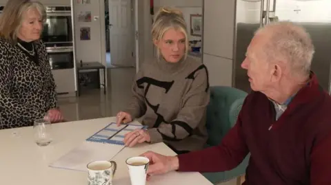 A woman with blonde hair sits in a kitchen, holding a calendar, talking to two elderly people either side of her. 