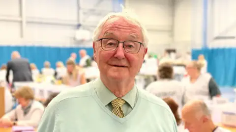 Councillor Roger Evans with white hair and wearing square black thin frame glasses. He is wearing a light sage green shirt and sage green jumper on top with a black and yellow tie tucked under the jumper. He is pictured inside a sports hall with a blue curtain around walls.