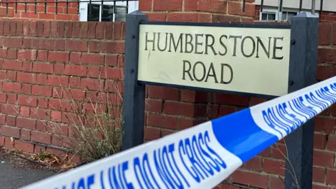 Eric Johnson/BBC A blue and white police tape which says POLICE LINE DO NOT CROSS, in the foreground. Behind is a road sign reading HUMBERSTONE ROAD, and a brick wall.