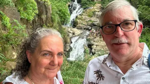 Alan Dutton A couple in their 60s stand in front of a waterfall which has rocks and greenery around it. She is wearing a white shirt and gold chain necklace, her hair is shoulder length and clipped back from her face, and is grey and curly and she has the strap of a brown bag across her shoulder.   He has white hair and a moustache and is wearing a white shirt with a palm tree pattern.