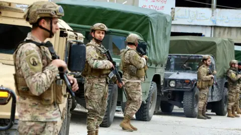 Five Lebanese army soldiers stand with guns in front of three army trucks.