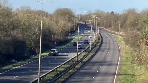 A carriageway pictured from a bridge. On the left side vehicles are travelling away from the camera. On the right handside the road is empty. There is a grass verge on the right and the road has a central reservation which has grass and railings. 