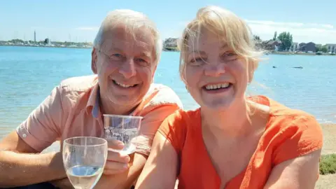 Chris and Ruth Stone-Houghton sit together with drinks outside a waterfront pub, smiling at the camera. He has white hair and wears a light-coloured, short-sleeved, collared shirt. She has short, blonde hair and wears an orange blouse.