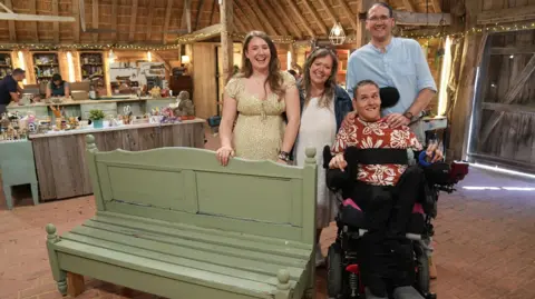 Freya, Madeleine, Andy and Ade (in a wheelchair) stand behind a green bench on the Repair Shop set