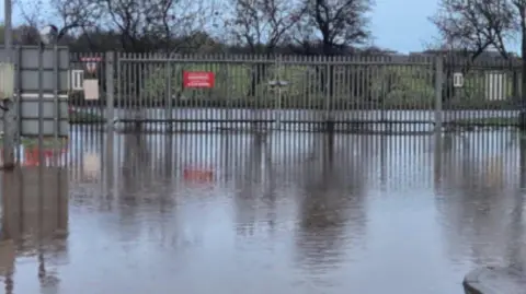 Westmorland and Furness Council Flusco household waste recycling centre in Penrith. There is deep standing water at the iron gates of the centre. It is a dark, gloomy day. 