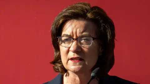 Eluned Morgan gesturing towards the camera, stood in front of a microphone. She is wearing glasses and is in front of a red background