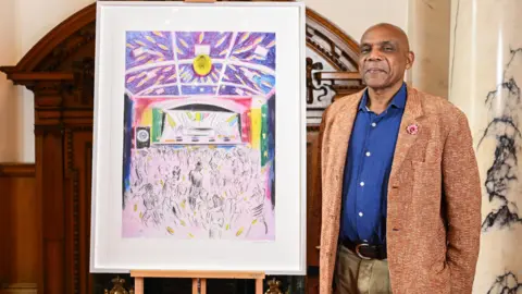 PA Media British artist Denzil Forrester - a man smiling, wearing a navy buttoned shirt, a brown tweed suit jacket and light brown trousers, standing to the right of his artwork depicting a reggae festival in Cornwall.