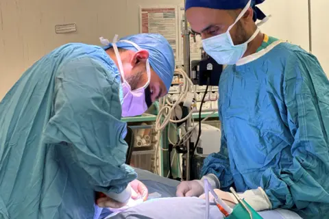 Newcastle Hospitals/PA Media Neurosurgeon Akbar Hussain (left) and Dr Sachin Rastogi operating on Ralph Seed. They are both wearing blue surgical gowns and facemasks.