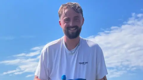 Ryan Cruikshanks Ryan Cruikshanks has short brown hair and a short brown beard. He stands in front of the blue sky with a cloud across it. He wears a white top with a "Half DRAM 2025" label and holds a bottle of Lucozade.