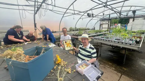 Five people sit around a table in a greenhouse. There is a blue box full of produce on the table in front of them, and a row of pot plant seedlings behind them.