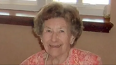 Una Crown smiles at the camera. She has brown hair and is wearing what looks like a pearl necklace with matching earrings. She is wearing an orange dress with a white flower pattern. Part of a window can be seen behind her.