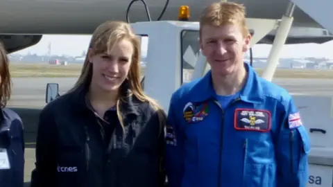 Susan Charlesworth Susan Charlesworth has long blonde hair and is wearing a green jumpsuit. She is standing next to Tim Peake, who has a blue jumpsuit on and short ginger hair. Behind them is a runway and a plane.