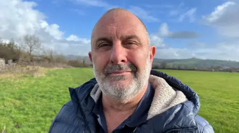 Man with beard in blue puffer coat standing in field beneath blue sky.