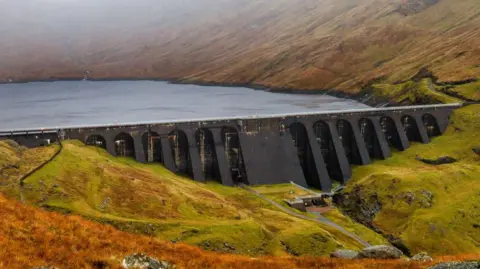 The dam is dark grey in colour with the grey waters of a reservoir behind it. The dam is in a mountain landscape of heather-covered hills.