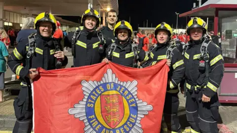 Jersey Fire Service Five Jersey Fire Service crew members in full kit, and another crew member not in full uniform, hold a flag with the service's yellow red and blue badge on it before starting the Around the Island Walk.
