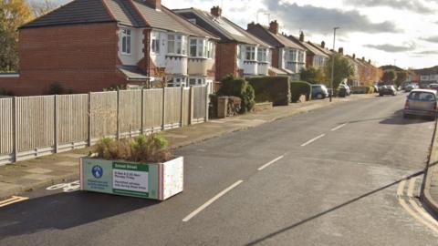 A general view of Northfold Road in Knighton, Leicester, with a planter in the middle of the left lane, restricting access to it. The planter has a sign which has rules about prohibiting drivers during school run times. 