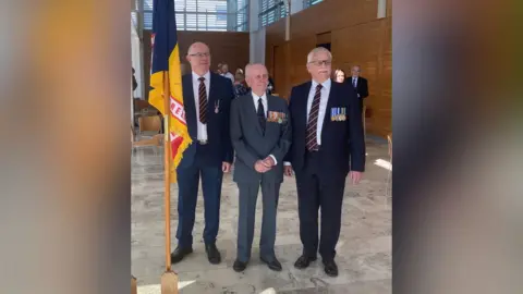 Norman Irwin Three men stand looking off camera. They all wear suits and have various military medals.