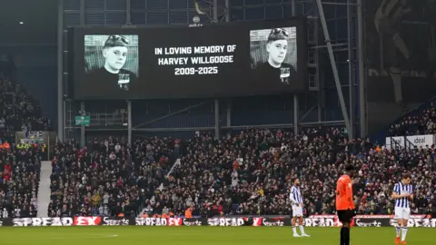 PA Media A screen at The Hawthorns football stadium reading 'in loving memory of Harvey Willgoose 2009-2025' with fans beneath it. Three players can be seen on the pitch.