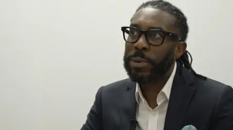 Teacher Marlon James-Edward, wearing a dark jacket and pale shirt, an black-rimmed glasses. He is on the right of the image, pictured mid-sentence with his mouth slightly ajar. He looks up toward the top left of the image as if thinking.