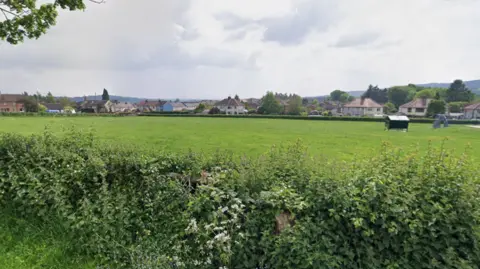 A large green field of a recreation area with a hedge in the foreground and houses beyond it. To the right you can see part of a play park