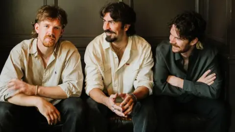 Adam Heffernan Amble - Oisín McCaffrey (a medium-length ginger haired man with a cream shirt and jeans), Robbie Cunningham (medium-length brown hair and beard, with a white shirt and jeans) and Ross McNerney (also medium length brown hair, a moustache and dark jacket) - all pictured from the waist up, sat own a bench. Oisin is staring at the camera intensely, Ross and Robbie are staring at him and smiling.