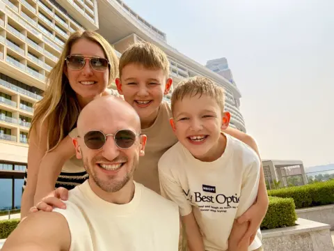 Adam Callow poses for a selfie photo with his wife and two young songs, with high rise flats and blue sunlit sky in the background.