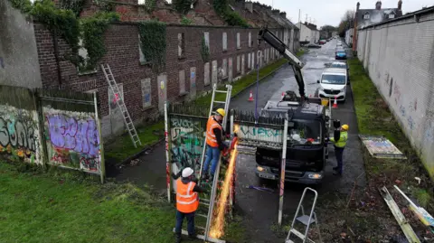 Three men in Hi-Viz jackets dismantle a steel wall that has been covered in multi-coloured graffiti. The wall sits at the end of a street with derelict red-brick houses running perpendicular to it. One of the men is on-top of a ladder with a steel cutter in hand. Sparks can be seen flying of the cutter. 