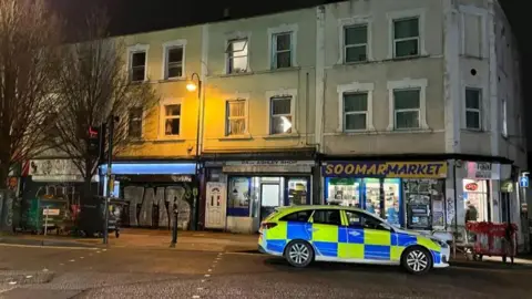 A police car, pictured to the bottom right of the image, parked on a street after dark with the road lit by a lamp-post. Shop fronts can be seen on the ground floor of a row of buildings with windows on the floors above and trees to the left of the image.