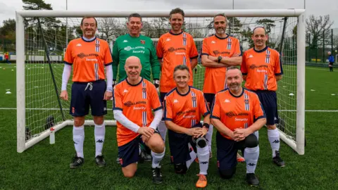 Seven men in orange football shirts and dark blue shorts and one man in a green football shirt standing on a grassy football pitch with goal posts behind them