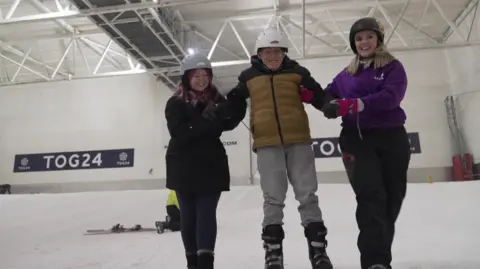 A man smiles as he skis down an indoor snow slope held by two women on either side of him. The man is wearing a brown and black jacket, grey jeans, black boots and a white helmet. The woman on the left has purple hair and is wearing a grey helmet, black coat and dark leggings. The woman on the right has blond hair and is wearing a black helmet, purple jacket and black trousers.