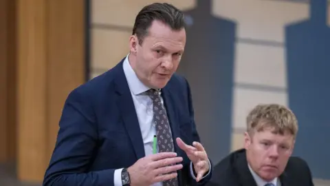 PA Media Russell Findlay, who has dark hair slicked back, speaks with his hands held out in front of him. He is wearing a blue suit and shirt with a patterned tie. 