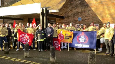 Image of a group of firefighters standing with banners and flags outside a red brick building.