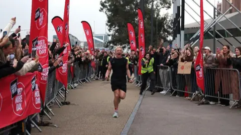 Getty Images Jamie Laing running through a marathon finish line while wearing a black tank top and shorts. He is clenching his fists and shouting with joy 