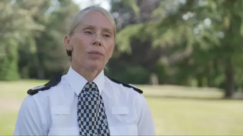 Assistant Chief Constable Lynn Ratcliff in a white police uniform shirt with black epaulettes and a black-and-white checkered tie, standing outdoors in a park-like setting. Trees and grass are visible in the softly blurred background, and the image is framed as a head-and-shoulders portrait.