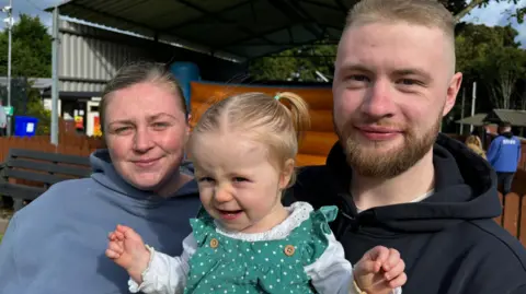 A mother, father and their daughter smile at the camera while attending the fun park at Carnfunnock Country Park. Jennifer is wearing a blue hooded top, their young daughter is earing a white top and green dress and her hair is in pigtails. Her dad has blond hair and a beard and is wearing a black hooded top