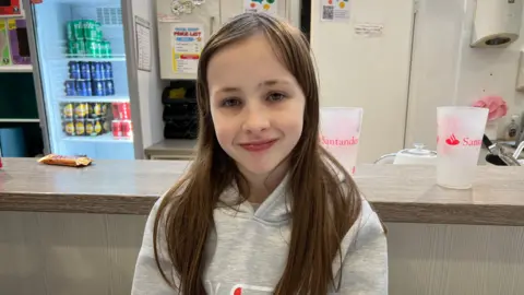 BBC Mia has long brown hair and is wearing a grey hoodie. She is standing in front of a counter at the centre.