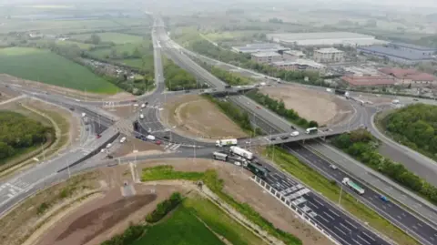 Moto Hospitality Limited A drone image of the roundabout which will allow both eastbound and westbound access to the new service station just off junction 16 of the M4 in Swindon, with lots of traffic on the roads, and large buildings and greenery in the background. 