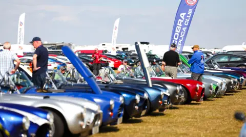 Silverstone A row of classic racing cars on display at an event, with enthusiasts looking around at the vehicles on display.