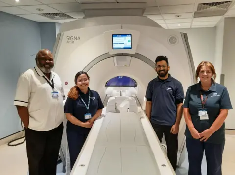 Four members of staff stand around an MRI imaging machine. The trolley where a patient would lie, is outside the actual machine, which has a small circular entrance.
