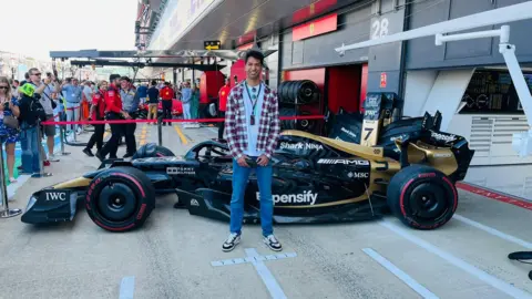 Supplied Man in a checked shirt and jeans standing in front of a racing car