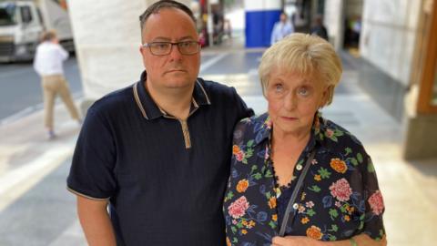 Kevin Hogg, a man with glasses, wraps his arm around Ann Ming, who is wearing a floral blouse and has blonde hair.