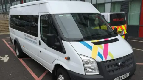 Wessex Cancer Support A white Ford Transit minibus with Wessex Cancer Support branding parked outside a hospital, with an ambulance beside it.
