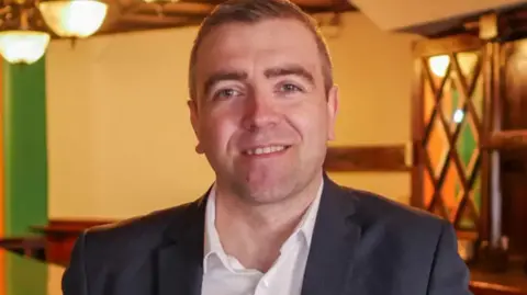 Steven Crosbie A man with short brown hair in a black blazer and white shirt looks directly at camera while vaguely smiling.