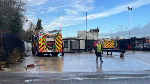 Two fire engines are parked at the entrance to an industrial unit. A man wearing a hi-vis vest can be seen walking towards the road. A second person stands next to the fire engine on the left.